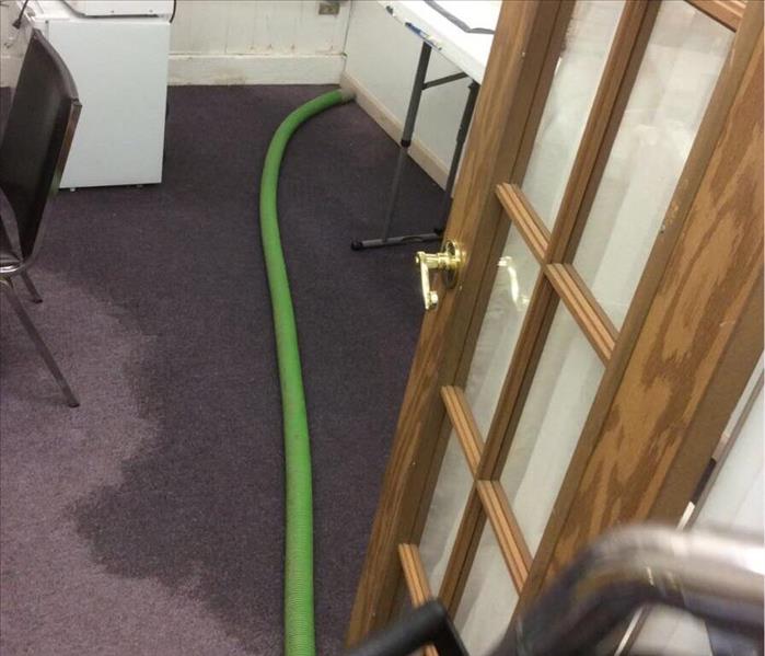 Blue carpet in room with wet spot along the wall with green hose and mini fridge and microwave 